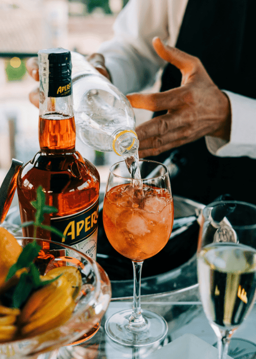 Aperol Spritz wird mit Mineralwasser und Eiswürfeln in ein Weinglas eingeschenkt