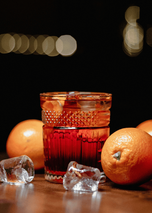 Roter Aperitif mit Eiswürfeln im Kristallglas, umgeben von frischen Orangen