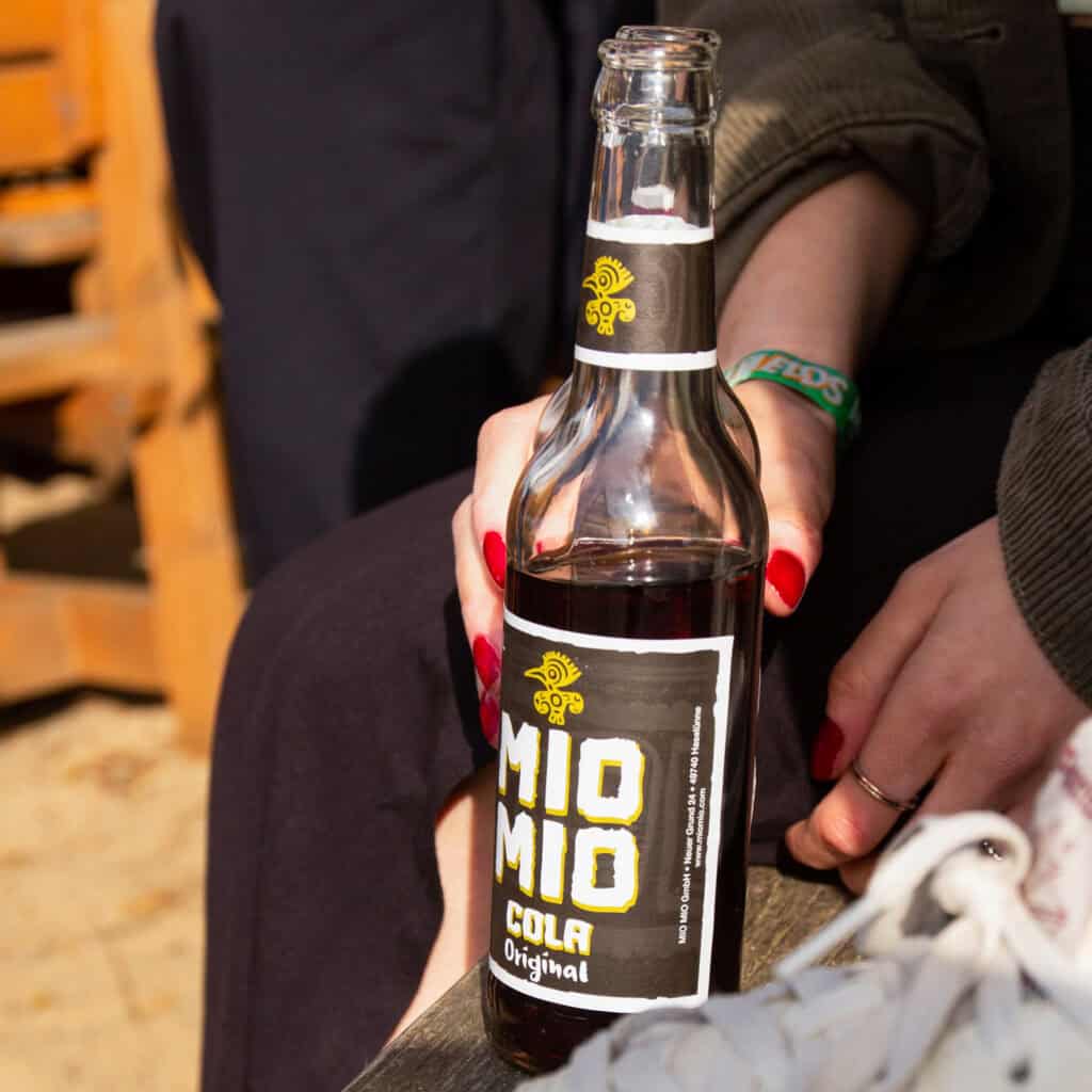 Mio Mio Cola Original in Glasflasche auf einem Festivalgelände in der Hand einer Besucherin